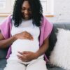 Cheerful pregnant African American female touching tummy gently while sitting on comfortable sofa with pillows in light room at home