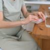 Unrecognizable pregnant female sitting at wooden table with glass of water while taking prenatal vitamins in light kitchen at home