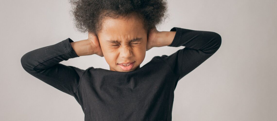Unhappy African American girl with Afro hairstyle covering ears while standing on white background with closed eyes in light studio