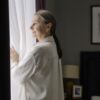 A senior woman with grey hair stands by a window, deep in thought, in a cozy home setting.