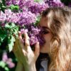 A young woman relishes the scent of lilac flowers in a sunlit garden, embodying spring and relaxation.