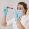 A female healthcare worker wearing gloves and a mask examines a blood sample in a test tube.