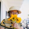An elderly woman embraces nature, smelling bright yellow flowers on a sunny day.