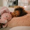 A young girl sleeping peacefully with a pink stuffed animal in a cozy bedroom environment.