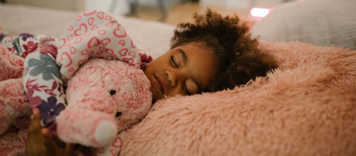 A young girl sleeping peacefully with a pink stuffed animal in a cozy bedroom environment.