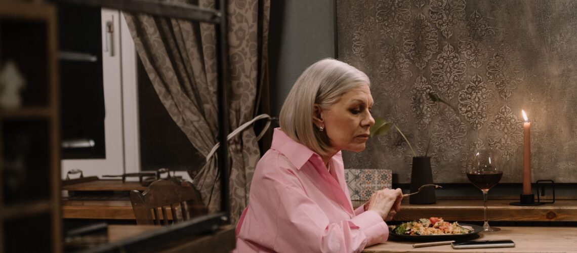 Elderly woman in a pink shirt enjoying dinner alone by candlelight at home.