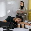 Exhausted employee resting on desk surrounded by documents, colleague in background.