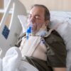 An elderly man lies in a hospital bed wearing an oxygen mask for medical care.