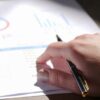 A close-up view of a hand holding a pen and examining financial charts on paper.