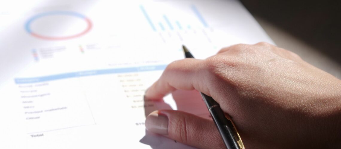 A close-up view of a hand holding a pen and examining financial charts on paper.