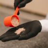 A close-up shot of a black-gloved hand pouring powder from a red scoop, over a textured surface.