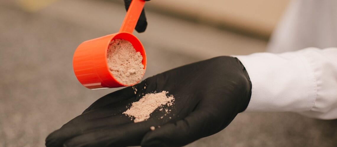 A close-up shot of a black-gloved hand pouring powder from a red scoop, over a textured surface.