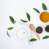 From above of cup with green tea near dry and fresh leaves with ceramic teapot on white table