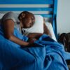 A woman rests peacefully in a hostel bed with a blue blanket and a backpack nearby, conveying travel fatigue.