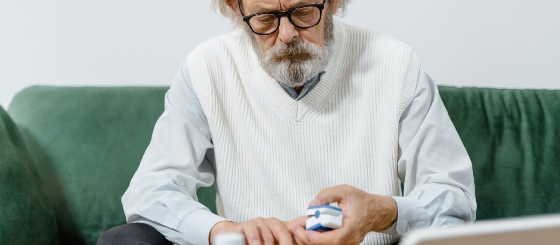 Elderly man with eyeglasses carefully examining his medication while seated on a green couch.
