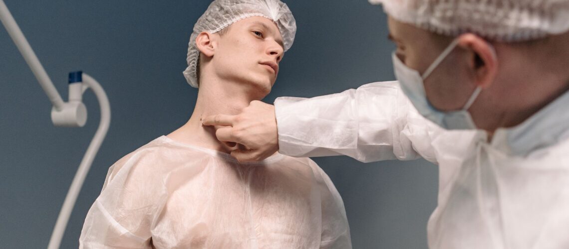 A doctor in protective gear examines a patient's neck in a sterile medical setting.