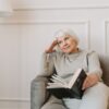 Senior woman with white hair relaxing indoors on couch, holding a book, appearing thoughtful.
