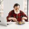 Elderly man in checkered shirt eating lunch at a table with a laptop and coffee indoors.