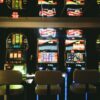 Slot machines in a dimly lit casino with reflective ceiling and empty chairs.