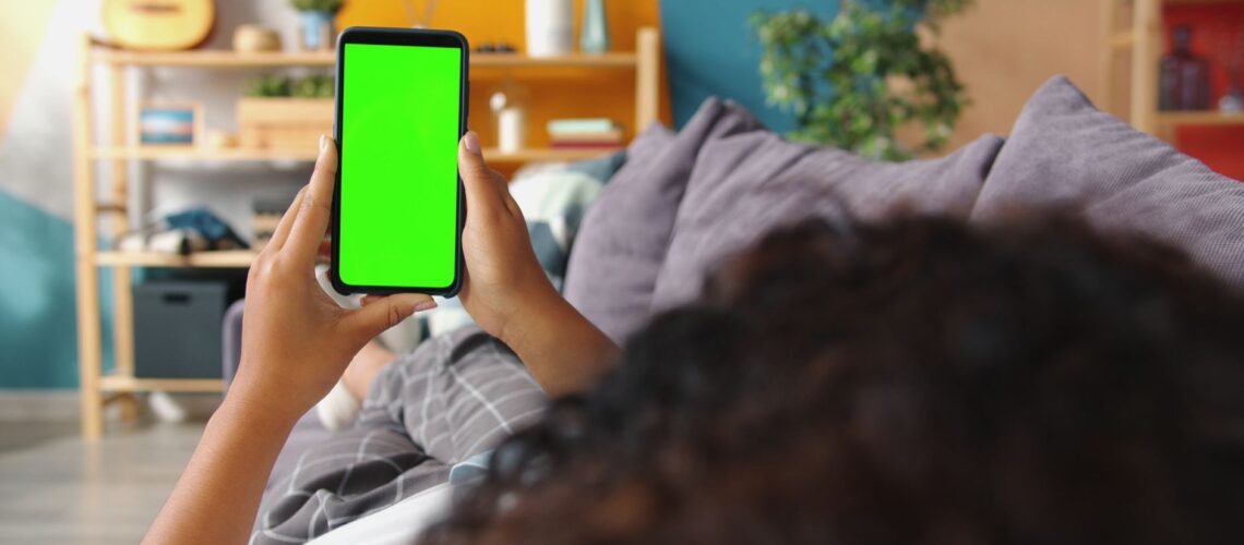 Person holding smartphone with green screen while relaxing on a couch indoors.