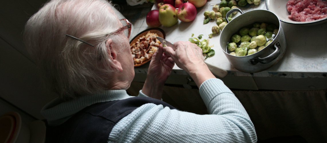 An elderly woman peels vegetables in a cozy kitchen setting, surrounded by fresh produce.