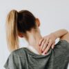 Back view of slim blond female in casual wear with manicure indicating pain spot on back while sitting in front of white wall with paper drawing in clinic
