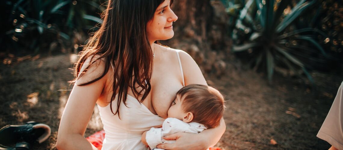 A serene scene of a mother breastfeeding her baby while sitting on a blanket outdoors in summer.