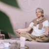 Elderly woman sitting on a sofa meditating with eyes closed, in a peaceful home setting.