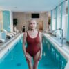 Senior woman in a red swimsuit stands confidently by an indoor pool, exemplifying grace and fitness.