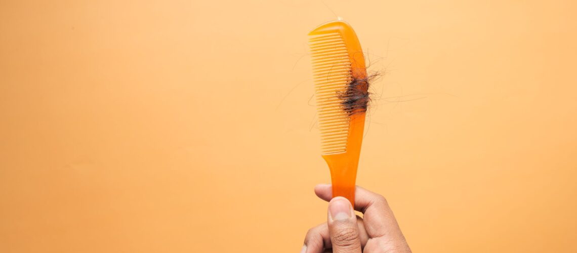 Close-up of hand holding a comb with hair against an orange backdrop, highlighting hair loss concept.