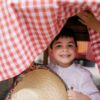 A cute child smiling while hiding under a red checkered tablecloth holding a straw hat.