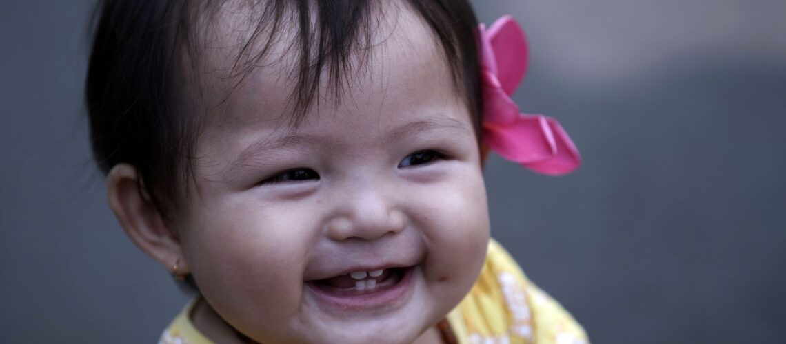 Smiling baby girl with pink hair bow and yellow dress, capturing pure joy.