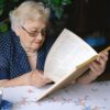 Serious female pensioner in eyeglasses reading book while sitting at table with cup of hot drink in room on blurred background