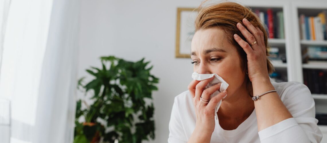 An adult woman feeling unwell, blowing her nose indoors.