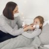 Mother administering nose drops to her sick daughter in a cozy bedroom setting.