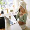 Pregnant woman in casual setting, talking on phone with laptop open and a drink nearby.