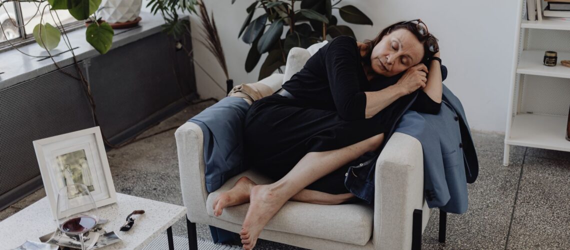A senior woman asleep on an armchair in a cozy indoor setting, surrounded by plants and a table with a glass.