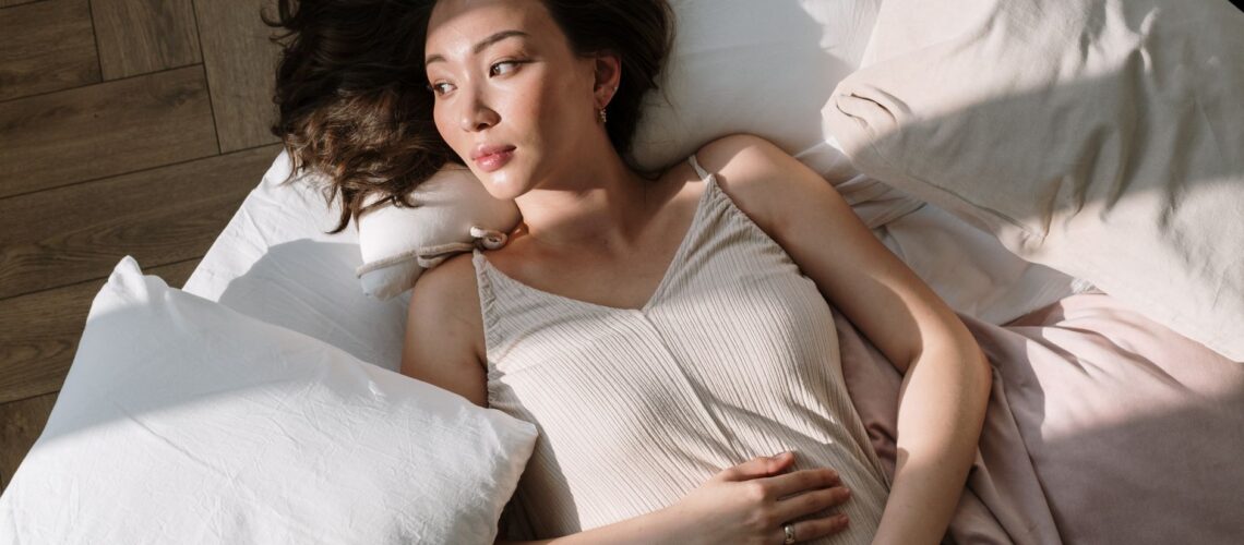 Asian woman in sleepwear lying on bed with hand on belly, enjoying morning sunlight.