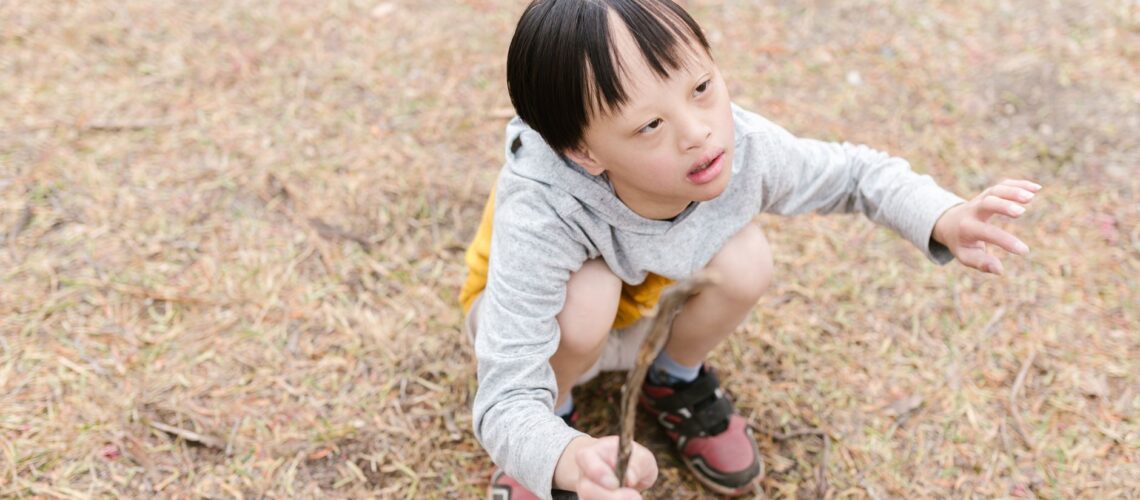 Young boy with Down syndrome enjoying outdoor play, crouching and holding a stick.