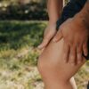 A close-up image of a person holding their knee, indicating pain or injury, with greenery in the background.