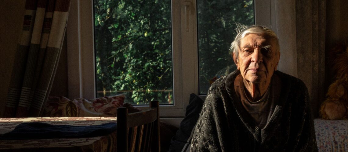 An elderly man sitting alone in a cozy room lit by sunlight through a window.