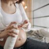 A pregnant woman applying lotion to her belly while sitting indoors wearing casual clothes.