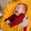 Emotional portrait of a crying newborn being comforted in caregiver's arms indoors.