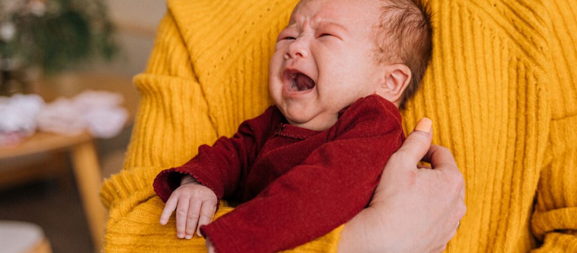 Emotional portrait of a crying newborn being comforted in caregiver's arms indoors.