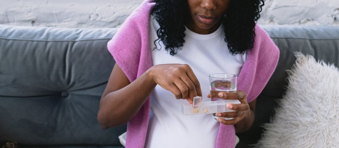 Serious pregnant African American female with glass of water taking prenatal vitamins while sitting on comfortable sofa in living room