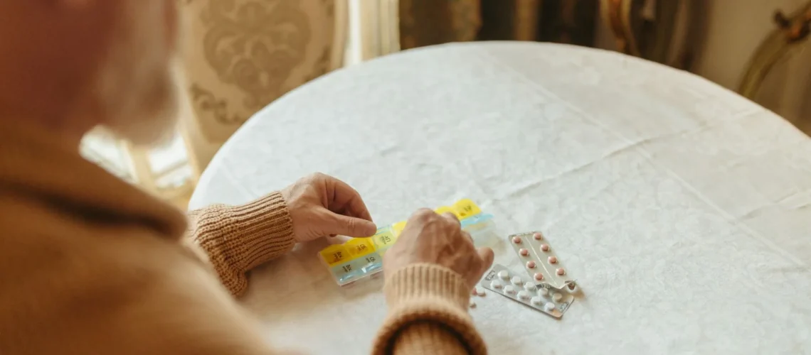 Personne âgée triant ses médicaments quotidiens avec un pilulier sur une table à l'intérieur.