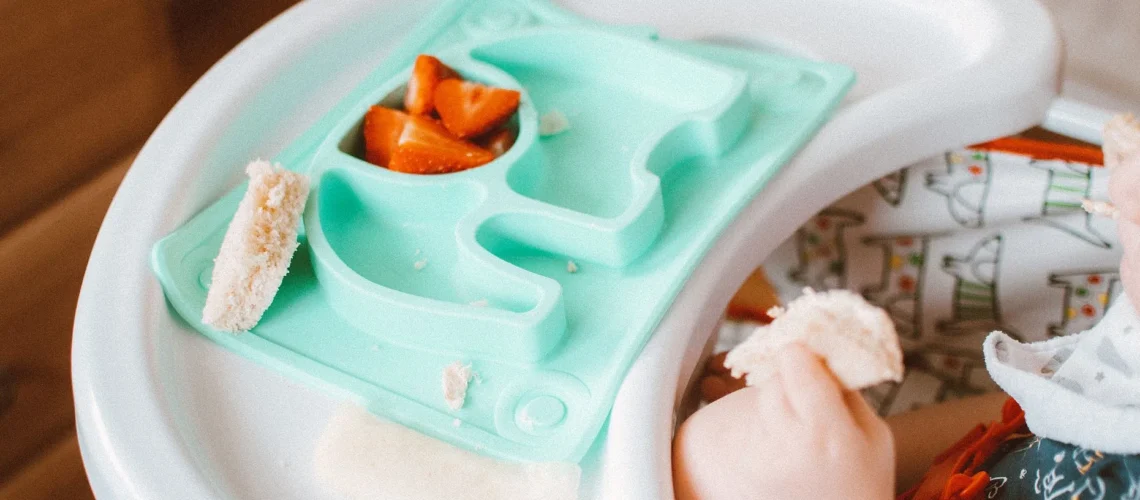 Bébé adorable mangeant du pain et des carottes dans une chaise haute avec un plateau coloré.