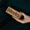 Main de femme tenant un bloc en bois avec le mot « Weeks » sur fond vert
