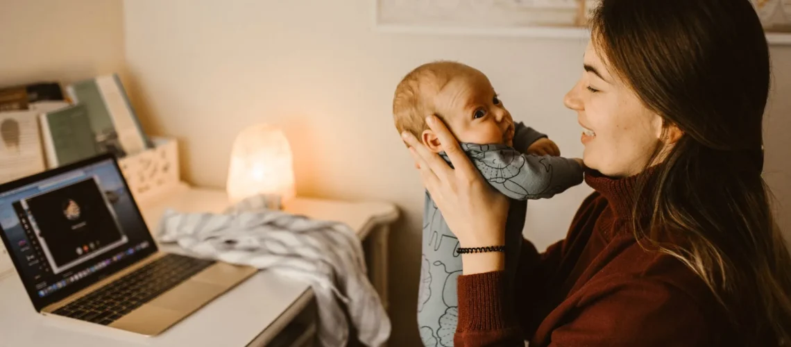 Mère tenant son bébé tout en passant un appel vidéo depuis son bureau à domicile. Scène intérieure chaleureuse et réconfortante.