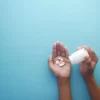 Mains versant des comprimés d'un flacon sur fond bleu, représentant soins et médication.